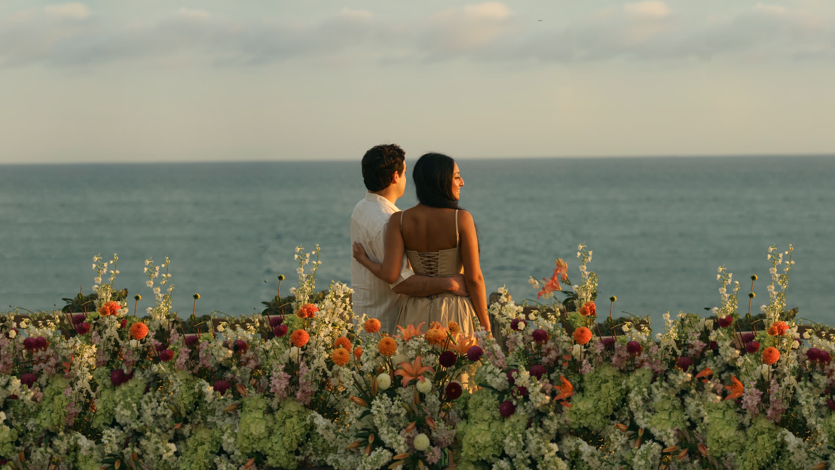 Naomi and Daniel looking out at the ocean behind a wall of flowers